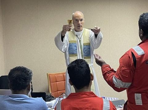 Baltimore ministry to ship's crew. Baltimore Auxiliary Bishop Parker raises Eucharistic elements as he says Mass in ship's hull. Stranded sailors pictured from behind, facing Parker.