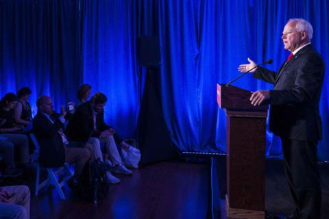 Minnesota Gov. Tim Walz, 2024. Gov. Walz pictured in profile, standing at lectern speaking with right hand raised in gesture. Room is covered in blue pipe and drape.