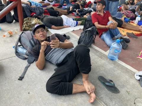 Migrants in caravan, Mapastepec, Mexico. Luis lies on the ground, using his backpack as a pillow, looking at his phone. Around him filling the frame are many other migrants.