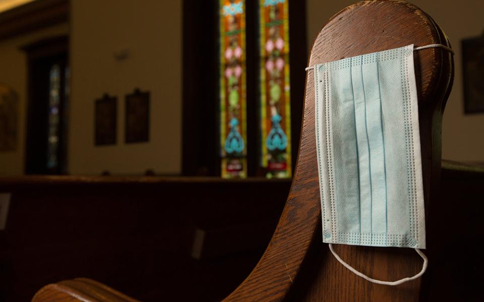 In this illustration photo, a disposable medical mask hangs on the side of a church pew. (CNS/ The Catholic Spirit/Dave Hrbacek)