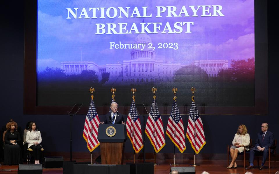 Biden stands behind a podium in front of several American flags and a projector screen that says "National Prayer Breakfast"