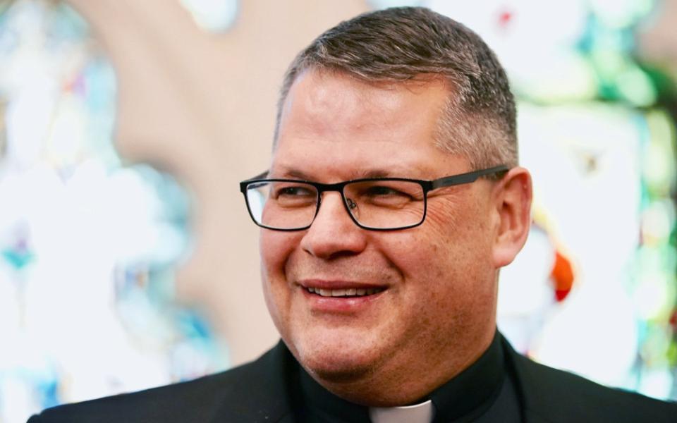 A white man with a short haircut wears glasses and a clerical collar and smiles.