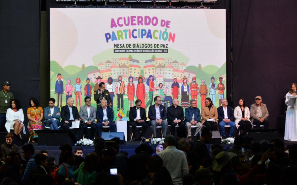Many people sit in a line under a screen with the words "Acuerdo de Participación" and colorfully illustrated images of people