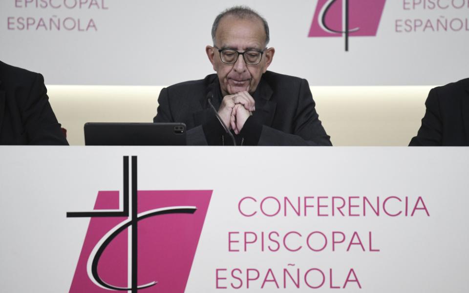 A white man wearing black and glasses closes his eyes and folds his hands as he sits at a table labeled "Conferencia Episcopal Española."