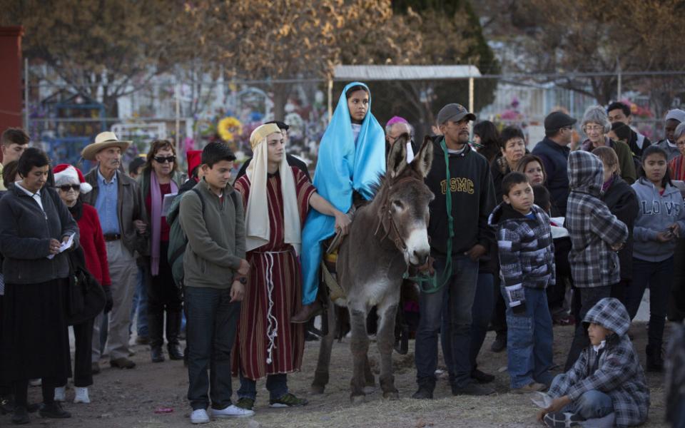 A crowd stands around a girl wearing a blue nativity costume riding on a donkey. A boy also dressed in nativity clothes stands beside her.