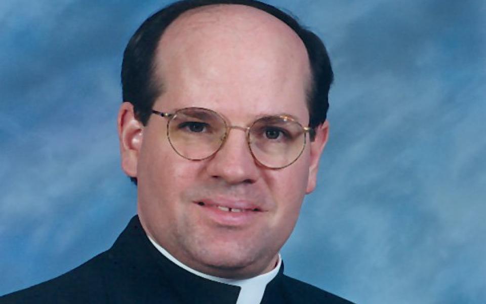 A balding white man with round glasses wears a clerical collar and looks into the camera