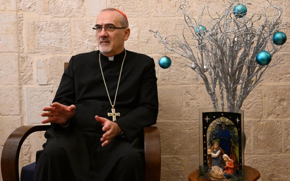 A man wearing a cassock, pectoral cross and red zucchetto sits next to a nativity scene and green orbs