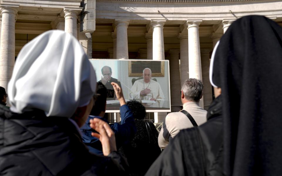 Pope Francis and a priest can be seen on a screen in front of a crowd with the backs of the heads of two religious sisters immediately in front of the camera