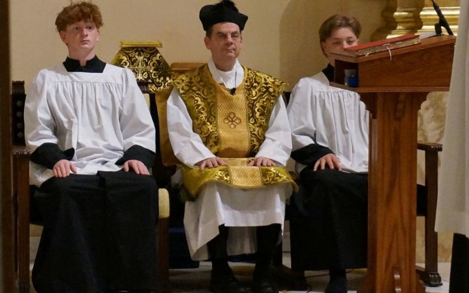 Fr. John Ubel, pastor of the Church of St. Agnes, wears the traditional biretta hat and vestments as he sits between altar servers during one of the Catholic parish's Latin Masses in St. Paul, Minn., June 28, 2026. (AP/Giovanna Dell'Orto)