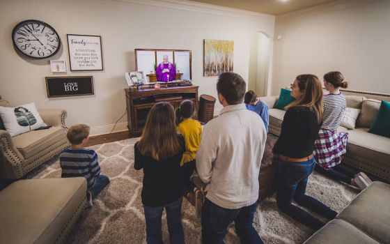 A family kneels in a living room while watching Mass livestreamed on TV