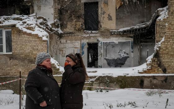 Two women stand outside a crumbling building in the snow. One woman cries.