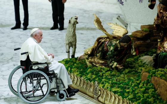 Pope Francis sits in a wheelchair in the snow facing a brightly painted creche