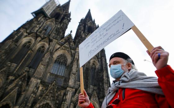 An older white man wearing a surgical mask carries a sign in the shadow of a cathedral