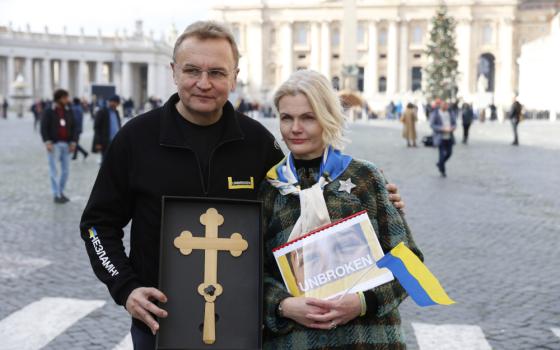 A white man with glasses holds a gold cross next to a white woman with a Ukrainian flag and a picture of a face with the words "unbroken" in St. Peter's Square