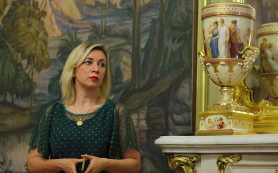 A blond woman with dark roots stands in front of a painting and ornate vase