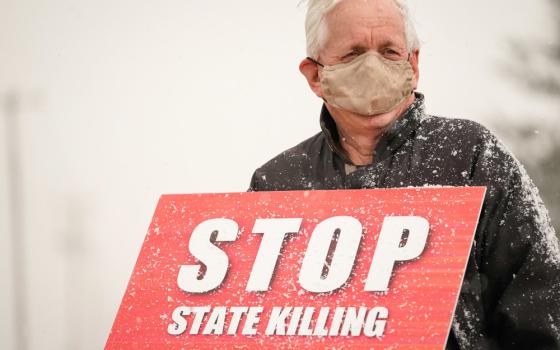 An older, masked man holds a sign that reads "Stop state killing" in the snow