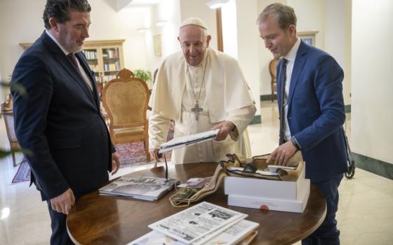 Pope Francis and two Spanish men stand around a table with newspapers and books on it
