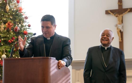 A Latino bishop speaks from behind a lectern. A Christmas tree, a crucifix, and a beaming Cardinal Wilton Gregory, an African-American man, stand behind him.