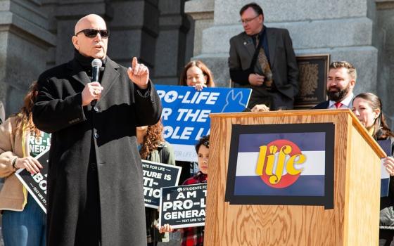 A bald man in black sunglasses and a long black coat holds a microphone and speaks in front of a crowd holding pro-life signs
