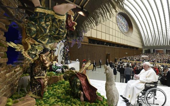 Pope Francis sits in a wheelchair in front of a large nativity scene indoors