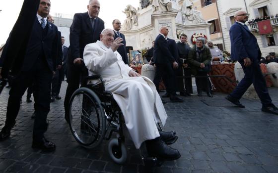 Pope Francis sits in a wheelchair pushed by a man in a suit and waves near a variety of statues