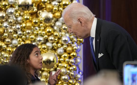 Biden leans down towards a girl with brown curly hair. They stand in front of silver and gold orbs