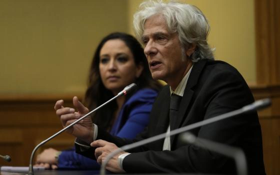 A man with floppy white hair sits next to a woman with brown hair behind microphones
