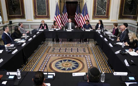 People sit at tables in a rectangle with Dough Emhoff at one end flanked by several American flags