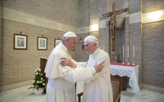 Pope Francis and Pope Benedict XVI, both in white cassocks, grip each others arms in a room with a crucifix and altar