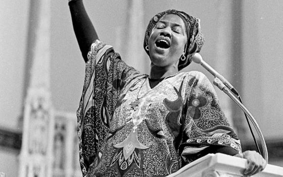 A black and white photo of a Black woman at a lecturn with one arm raised in the air. She wears patterned fabric around her head and torso.