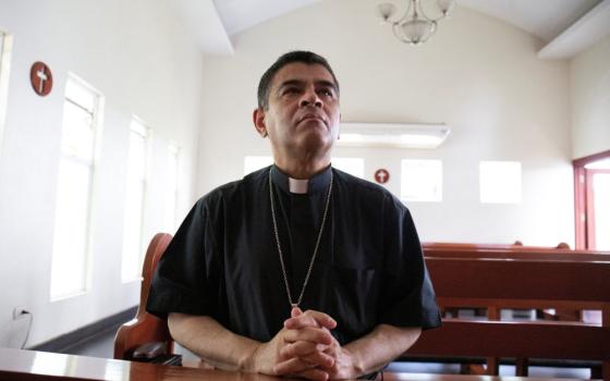 A light brown skinned man wearing a clerical shirt and a pectoral cross folds his hands in prayer