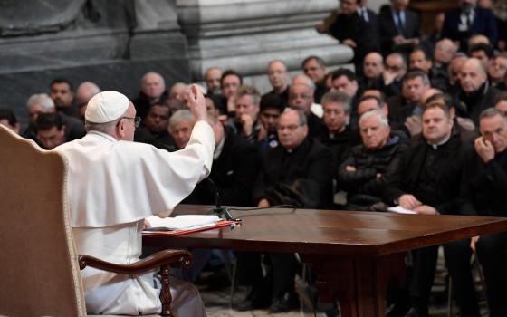 Pope Francis sits at a large table in front of a rows of many priests 