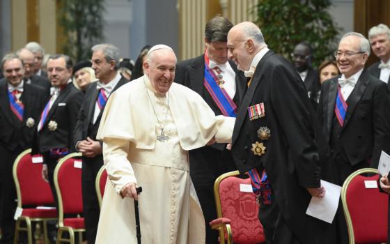 Pope Francis smiles broadly, holds a cane, and pats the arm of a white man wearing a tuxedo