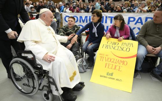 Pope Francis, sitting in a wheelchair, rolls by seated guests in front of a crowd