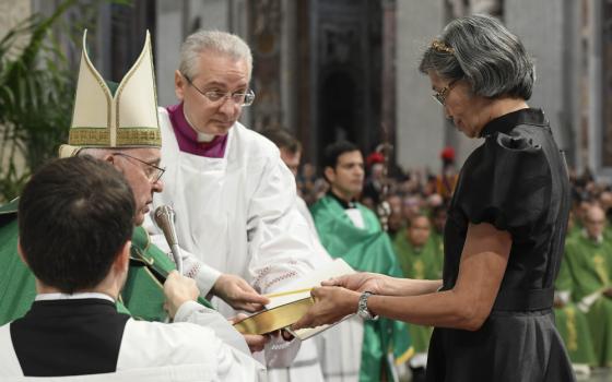 Pope Francis, who is sitting, hands a Bible to a standing woman with short gray hair and a black dress
