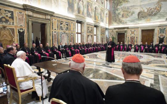 A bishop stands in the middle of an ornate room and speaks to Pope Francis and some cardinals