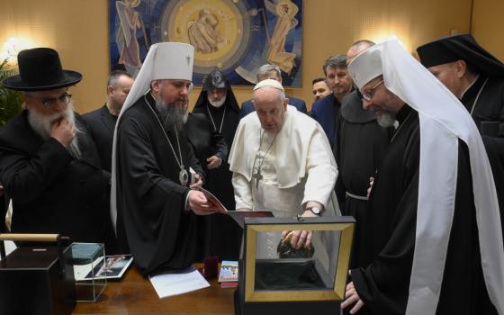 Pope Francis looks inside an open box with a crowd of older men in religious garb standing around him