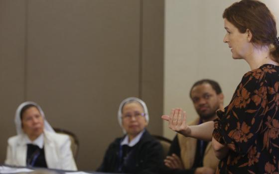 A white woman with curly brown hair stands in front of two seated women religious and a man, all people of color