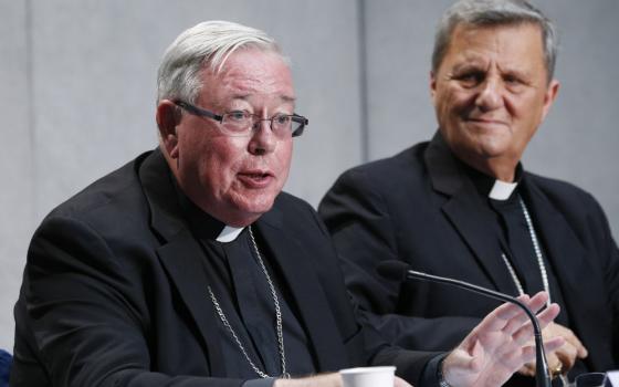 An older white man in a clergy collar and black suit and pectoral cross speaks into a microphone and is sitting next to another similarly dressed older white man