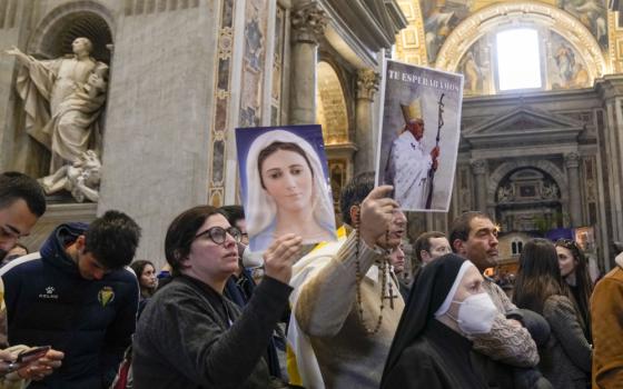 A man holds a rosary and a picture of Benedict that says "Te Esperabamos" in the middle of a crowded line