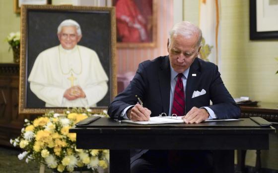 Biden sits at a desk and signs a book with a portrait of Pope Benedict XVI in the background