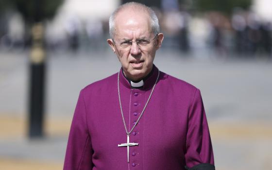 An older white man with glasses wears a magenta shirt, a clergy collar, and a cross necklace