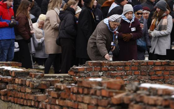 Two older wearing light blue and white striped handkerchiefs around their necks lay candles on a brick wall with a crowd behind them