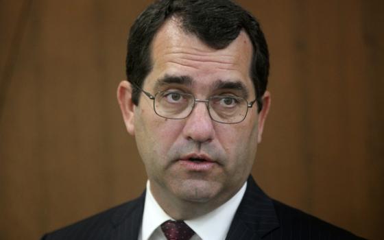 A headshot of pale-skinned man with brown hair and glasses wearing a suit and tie