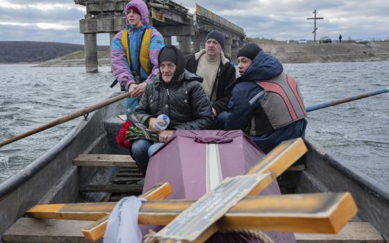 Four people with winter coats sit in a rowboat with a coffin and large cross
