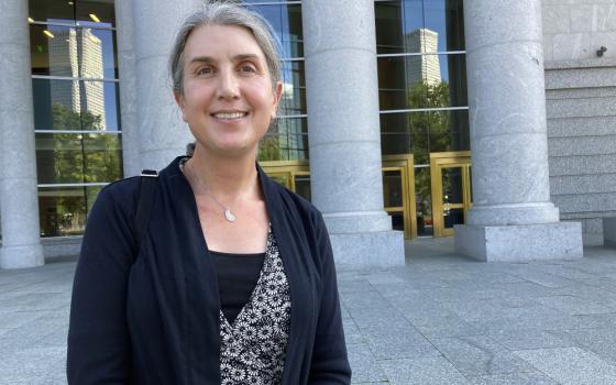 An older white woman wearing a cardigan stands outside a building with white marble columns