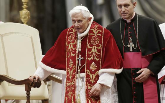 A white bishop stands behind Pope Benedict XVI who is wearing red and gold vestments