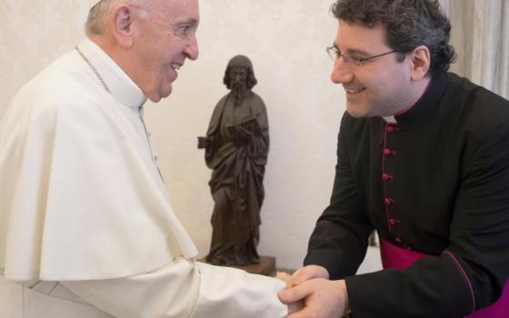Pope Francis shakes a man's hand who is wearing glasses and a black and purple cassock