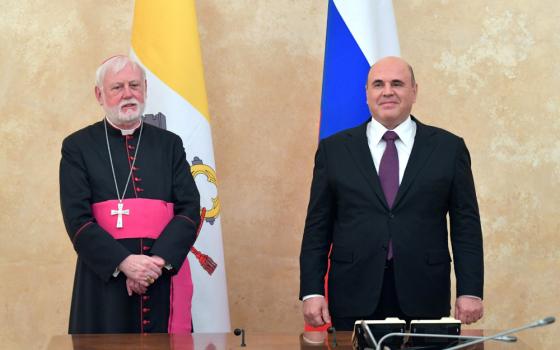 A man wearing a bishop's cassock stands next to another man wearing a suit and tie. A Vatican and Russian flag stand behind the two.