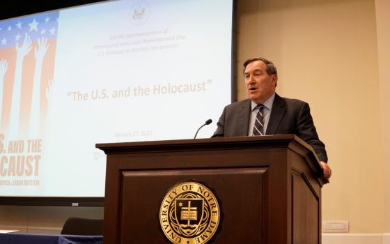A white man in a suit speaks from behind a podium with a projector and the words "The U.S. and the Holocaust" in the background